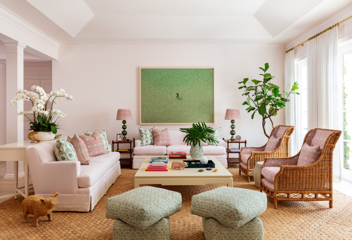 living room with pink walls and sofa, pink and green accents, 2 rattan chairs, 2 ottomans, and a fig tree in corner