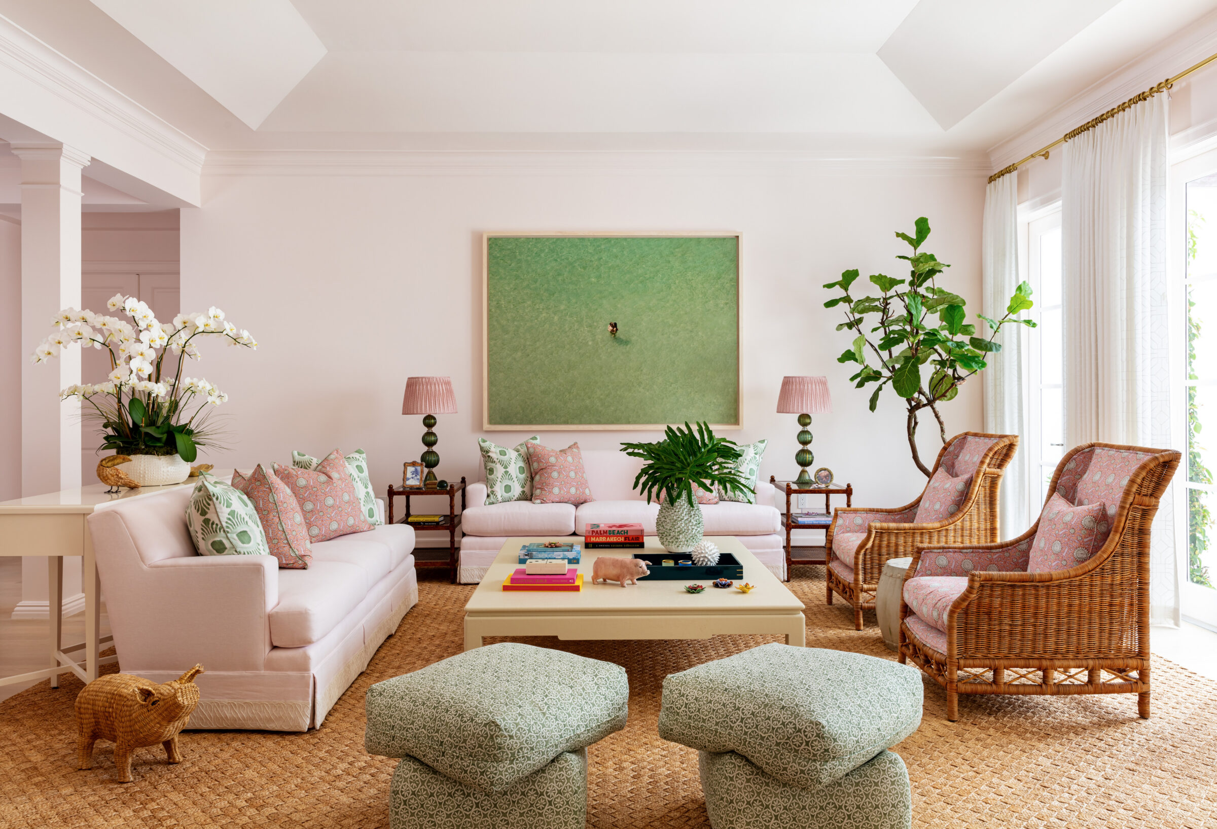living room with pink walls and sofa, pink and green accents, 2 rattan chairs, 2 ottomans, and a fig tree in corner