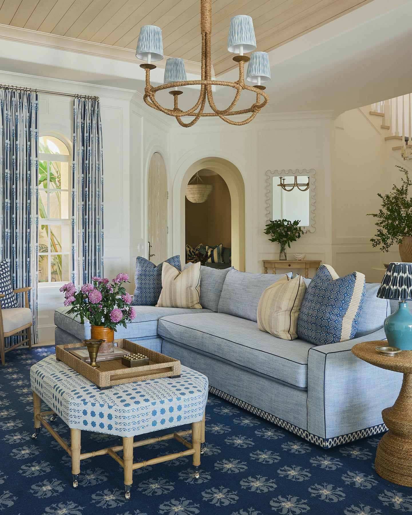 A living room showcasing a light blue sectional sofa, a dark blue patterned rug, an upholstered ottoman coffee table, and a rope-wrapped chandelier.