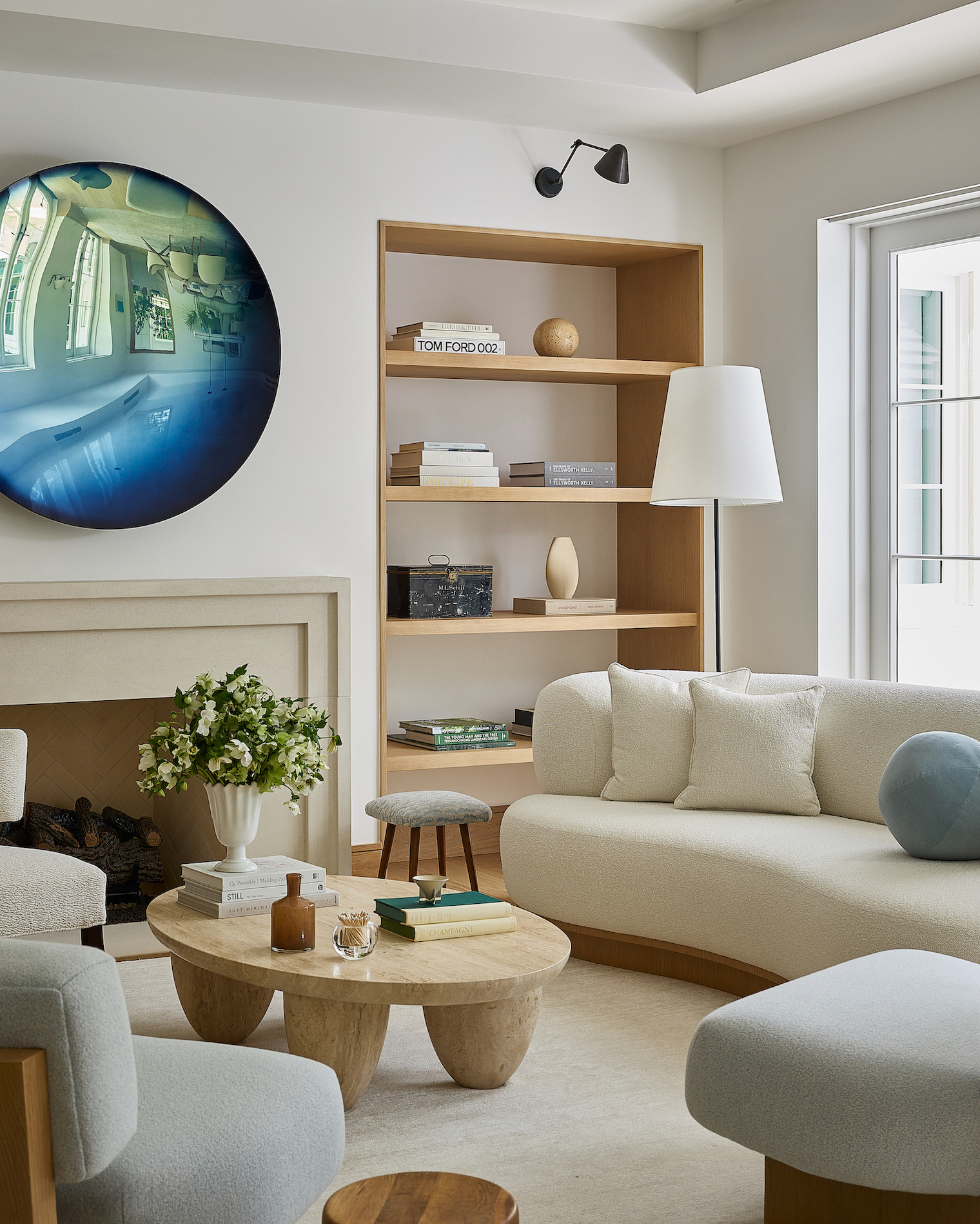 A bright living room corner featuring a stone fireplace topped with a round reflective blue art piece, surrounded by a curved cream sofa, blue lounge chairs, and a stone coffee table.