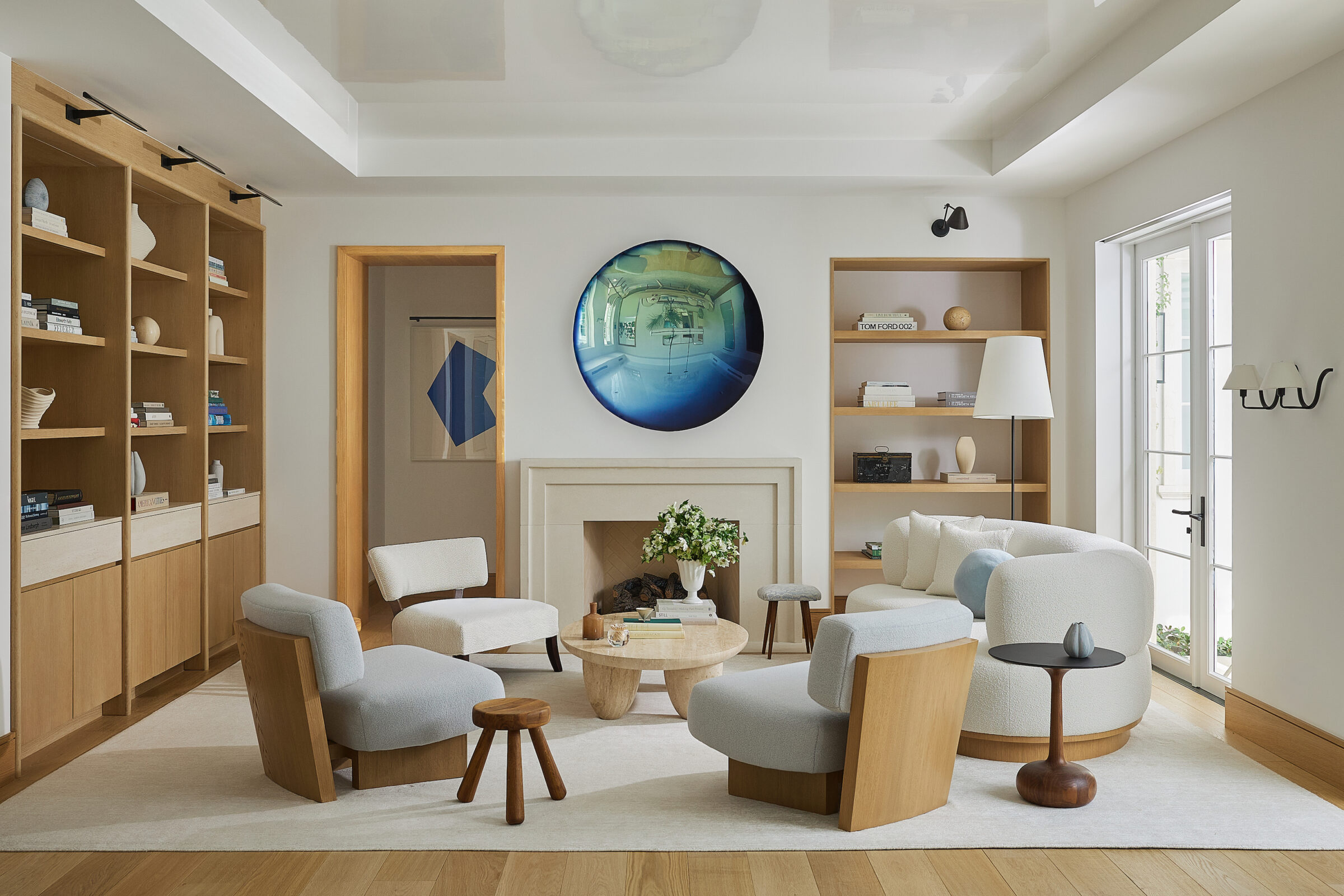 A wide view of a modern living room featuring a curved cream sofa, light blue lounge chairs, a stone fireplace with round blue artwork, and large wood built-in bookcases.